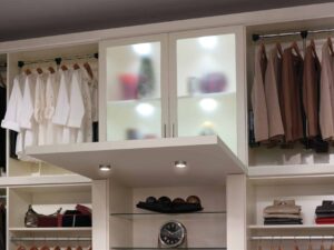 A neatly organized reach in closet features white shirts and brown jackets hanging, shelves with folded clothes, a clock, and a display cabinet with frosted glass doors. Closet Concepts of Grand Rapids