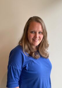 A woman with long, light brown hair wearing a blue shirt stands in front of a plain light-colored wall, smiling at the camera, ready to organize her space with new closet storage solutions. Closet Concepts of Grand Rapids