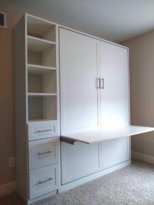 White Murphy bed with built-in shelves and drawers, shown partially unfolded with the bed frame extended in a carpeted room, offering versatile closet storage solutions. Closet Concepts of Grand Rapids