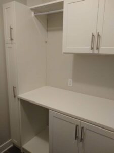 A white built-in cabinet setup for walk in closet storage, featuring upper and lower cabinets, a countertop, an open shelf, hanging rod, and a wall power outlet. Closet Concepts of Grand Rapids