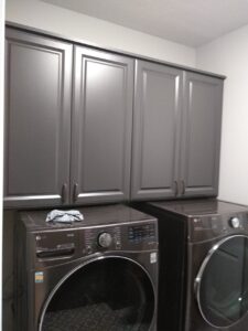 A laundry room with two front-loading washing machines below dark gray cabinets offers smart closet storage; a cloth rests on top of one machine. Closet Concepts of Grand Rapids