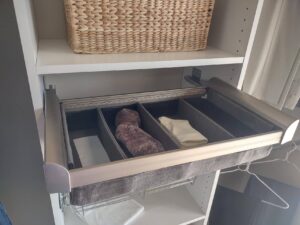 A closet drawer with divided compartments containing folded clothes and a pair of rolled socks, with a woven basket on the shelf above—perfect for maximizing reach in closet storage. Closet Concepts of Grand Rapids