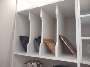 White vertical shelf dividers, often found in closet storage or closet systems, hold baking trays and cutting boards upright, while a pot is stored on a lower shelf. Closet Concepts of Grand Rapids