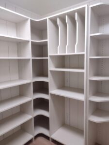 A white walk-in closet features multiple empty shelves, vertical dividers, and a corner shelving unit—highlighting smart closet storage solutions. Electrical outlets are visible on the back wall for added convenience. Closet Concepts of Grand Rapids