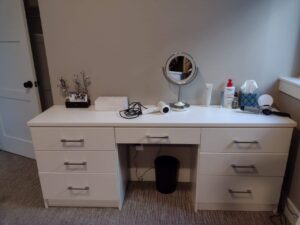 A white vanity desk with six drawers, a mirror, tissue box, jewelry stand, hairdryer, and toiletries on top is set against a beige wall—perfect for pairing with closet storage solutions. Closet Concepts of Grand Rapids