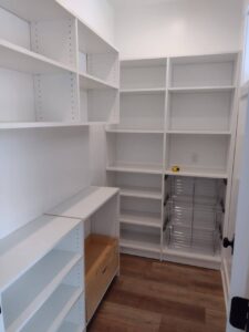 A walk-in pantry with empty white shelves, pull-out wire baskets, a wooden drawer, and a yellow tape measure on one shelf showcases versatile closet storage. The floor features medium-tone wood. Closet Concepts of Grand Rapids