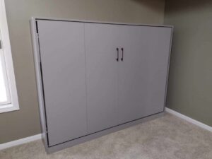 A closed gray murphy bed cabinet with double handles, positioned against a greenish wall on beige carpeted flooring, offers a smart space-saving solution inspired by modern closet concepts. Closet Concepts of Grand Rapids