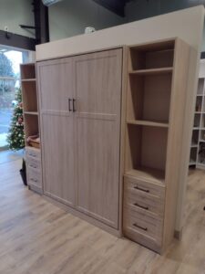 A light wood Murphy bed with closed cabinet doors, flanked by open shelves and drawers from versatile closet systems, is positioned in a showroom with a Christmas tree visible by the window. Closet Concepts of Grand Rapids
