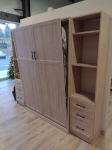 A wooden wardrobe with sliding doors, open to reveal clothes hanging inside, sits next to a shelving unit with drawers—showcasing stylish closet concepts. A Christmas tree is visible through a glass door in the background. Closet Concepts of Grand Rapids