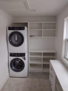 Laundry room with stacked washer and dryer on the left, open shelving and countertop on the right, closet systems for organized storage, and a window letting in natural light. Closet Concepts of Grand Rapids