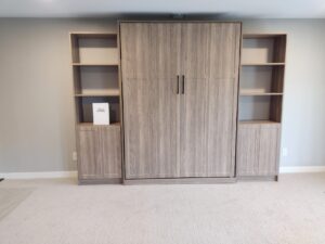 A wooden Murphy bed with closed doors is flanked by built-in shelves and cabinets, offering clever closet storage solutions against a light-colored wall in a carpeted room. Closet Concepts of Grand Rapids