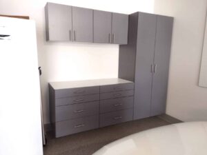 Gray storage cabinets with drawers and cupboards, ideal for closet storage, are installed against a white wall next to a refrigerator and beneath wall-mounted cabinets. Closet Concepts of Grand Rapids