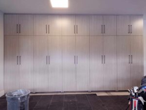A wall of light wood cabinets with black handles in a garage showcases smart closet concepts, with a trash bin and part of a golf bag visible in the foreground. Closet Concepts of Grand Rapids