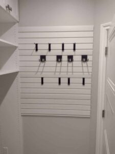 A white slatwall with multiple black hooks offers smart closet storage, mounted on a light gray wall in a closet beside a white door and open shelving. Closet Concepts of Grand Rapids