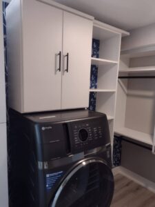 Front-loading washing machine beneath white storage cabinets and open shelving in a laundry room with gray flooring, patterned blue wallpaper accents, and clever closet systems for organized storage. Closet Concepts of Grand Rapids