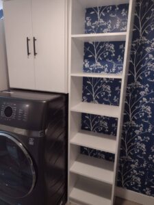 A laundry room with a washing machine, white cabinets above, and an empty white shelving unit offers excellent closet storage options against a blue floral-patterned wallpaper. Closet Concepts of Grand Rapids