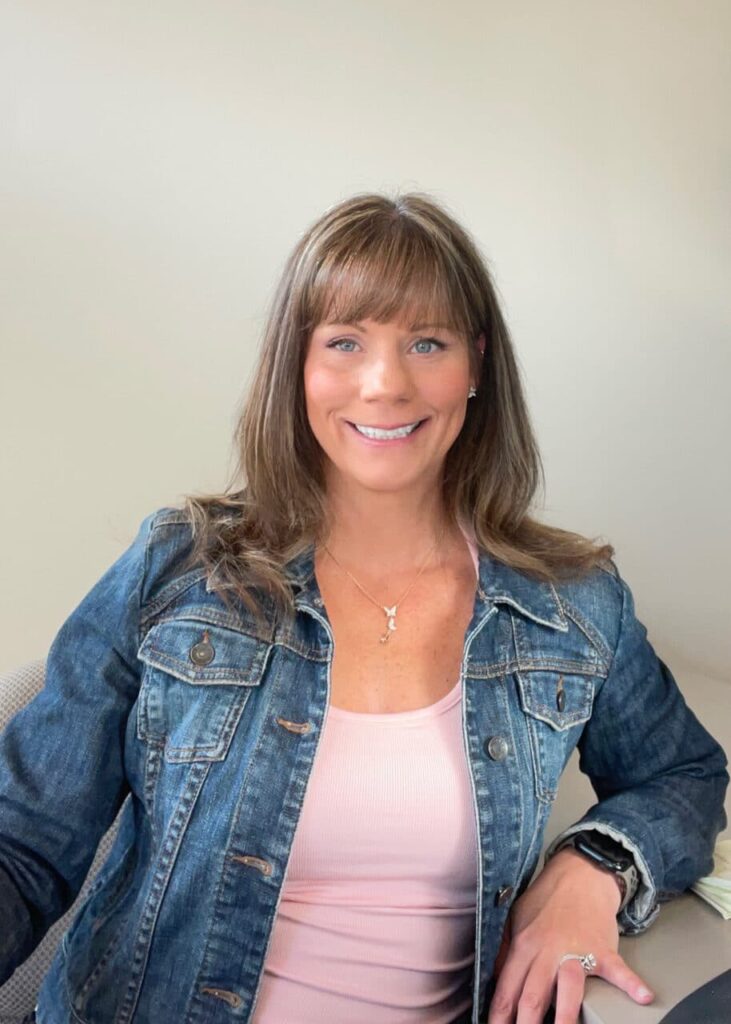 Woman with straight brown hair wearing a jean jacket and pink top, sitting and smiling at the camera in a neutral indoor setting, perhaps envisioning her dream walk-in closet or exploring modern closet systems. Closet Concepts of Grand Rapids