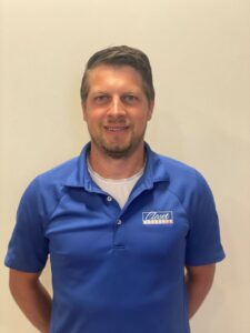 A man stands against a plain light-colored wall, wearing a blue polo shirt with a "Closet Masters" logo and a white undershirt, looking at the camera and smiling slightly—ready to help you with custom closet systems and storage solutions. Closet Concepts of Grand Rapids
