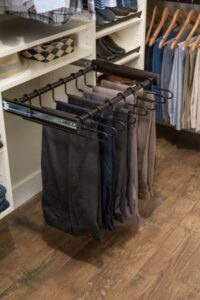 Pull-out pants rack with several pairs of pants hanging in a reach in closet, surrounded by shelves with folded clothes and shoes on a wooden floor. Closet Concepts of Grand Rapids