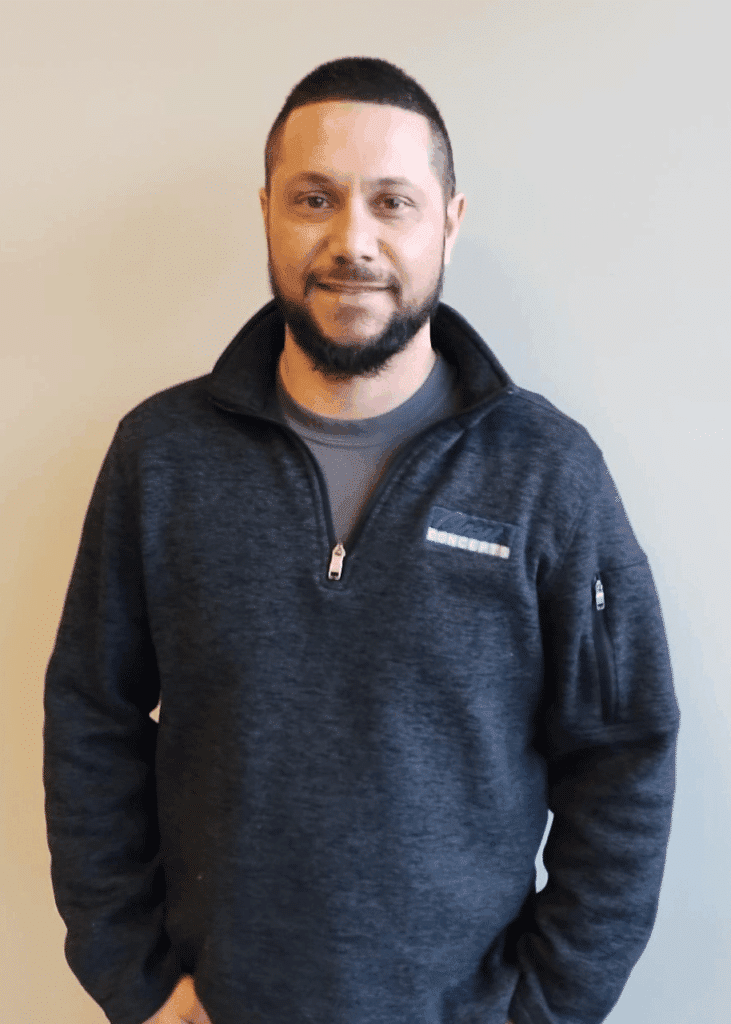 A man with short dark hair and a trimmed beard stands against a plain light-colored wall, wearing a dark zip-up sweater and gray shirt, as if considering closet storage solutions for his walk in closet. Closet Concepts of Grand Rapids
