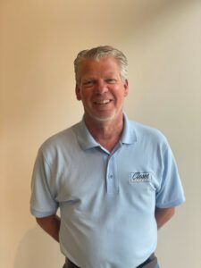 A man with gray hair wearing a light blue "Closet Concepts" polo shirt stands against a plain beige wall, smiling at the camera—ready to help you find the perfect closet storage or reach in closet solution. Closet Concepts of Grand Rapids