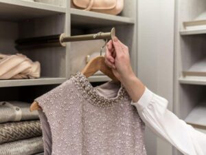 A person’s hand holding a beaded, sleeveless dress on a wooden hanger in front of an organized reach in closet with folded clothes and purses. Closet Concepts of Grand Rapids