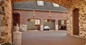 A brick house with three garage doors—one open to reveal a car inside. The foreground features stone columns, an arched entryway, and thoughtful closet concepts for organized reach-in closet storage throughout the home. Closet Concepts of Grand Rapids
