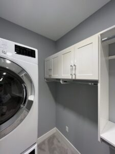 A modern laundry room with a front-loading washing machine, white cabinets, light gray walls, and built-in closet storage for added organization. Closet Concepts of Grand Rapids