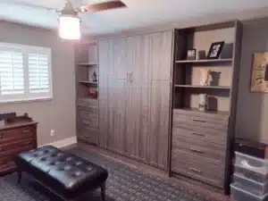 A wall unit with cabinets, drawers, and open shelves offers versatile closet storage in a room with a bench, dresser, and shuttered window. Closet Concepts of Grand Rapids