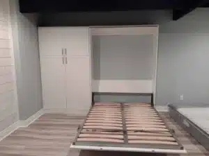 A white Murphy bed frame is pulled down from a wall cabinet in a minimally furnished room with light gray walls, wood flooring, and clever closet concepts for stylish storage. Closet Concepts of Grand Rapids