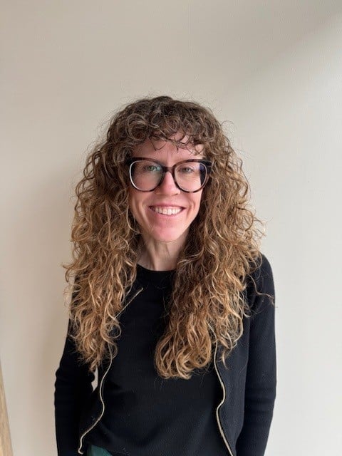 A person with long curly hair, glasses, and a black top stands smiling in front of a plain light-colored wall, ready to organize with smart closet systems. Closet Concepts of Grand Rapids