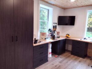 A workshop with dark wood cabinets, a reloading press on the counter, a wall-mounted TV, large windows letting in natural light, and smart closet storage for organized tools. Closet Concepts of Grand Rapids