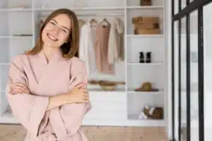 Woman in a light pink robe stands with arms crossed and smiles in front of an open wardrobe, showcasing organized closet storage and neatly arranged boxes. Closet Concepts of Grand Rapids