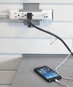 A power strip mounted on a wall has several cords plugged in, including a phone charging cable connected to a smartphone resting on a gray surface below. Closet Concepts of Grand Rapids