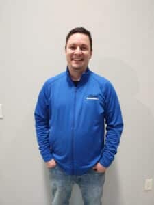 About: A man in a blue zip-up jacket and jeans stands against a light gray wall, smiling with his hands in his pockets. Closet Concepts of Grand Rapids