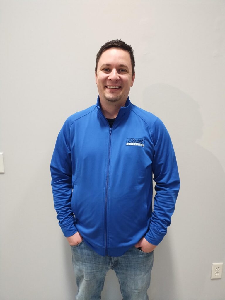 About: A man in a blue zip-up jacket and jeans stands against a light gray wall, smiling with his hands in his pockets. Closet Concepts of Grand Rapids