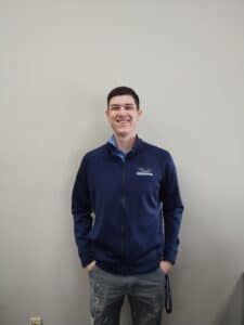 About: A young man wearing a navy blue zip-up jacket and jeans stands against a plain light-colored wall, smiling with his hands in his pockets. Closet Concepts of Grand Rapids
