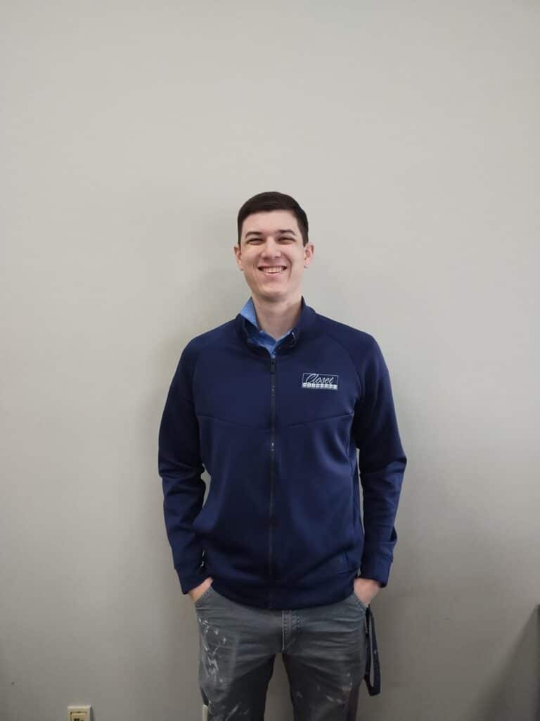 About: A young man wearing a navy blue zip-up jacket and jeans stands against a plain light-colored wall, smiling with his hands in his pockets. Closet Concepts of Grand Rapids
